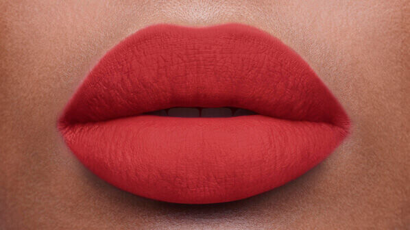 Close-up of a pair of full, matte red lips against a neutral skin tone background.