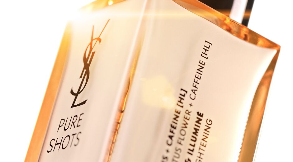 A close-up of a bottle of YSL Pure Shots skincare product, featuring a sleek design with a light orange liquid inside and a label that mentions ingredients like caffeine and lotus flower.