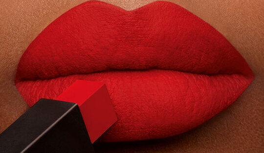 An extreme close-up of red lips applying a bold red lipstick, with the lipstick bullet prominently displayed.