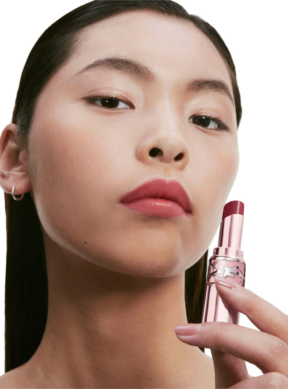 A close-up portrait of a model with straight, dark hair and minimal makeup, holding a lipstick in a pink tube. She has a confident expression and is looking directly at the camera.