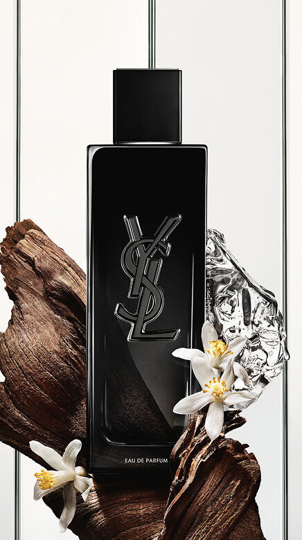 Une bouteille noire d'Eau de Parfum Yves Saint Laurent avec le logo YSL, entouree de morceaux de bois sombre, de fleurs blanches delicates et d'un objet reflechissant argente froisse.