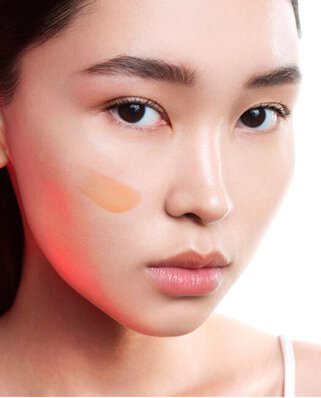 Nabijbeeld van een jonge vrouw met een lichte foundationstreek op haar wang, met neutrale make-up en een lichte achtergrond.