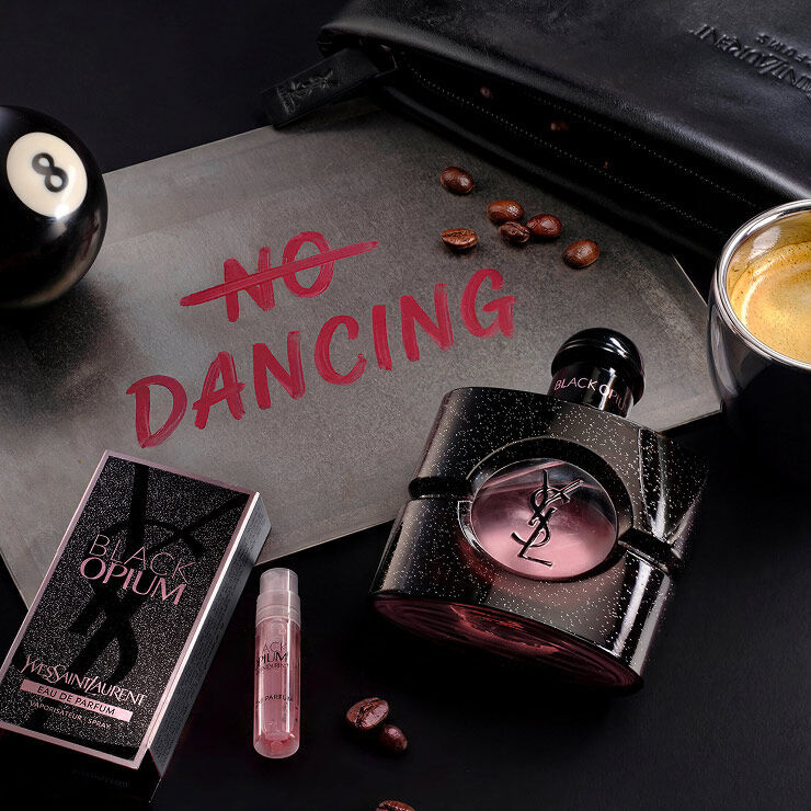 Black Opium Eau de Parfum and its sample vial next to a "No Dancing" sign, coffee beans, and a billiard ball.