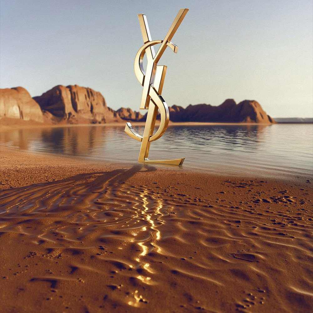 Un grand logo YSL (Yves Saint Laurent) dore en 3D pose sur un rivage desertique. Le monogramme dore poli reflete la lumiere du soleil et se trouve devant une etendue d'eau calme, avec des formations rocheuses rouges spectaculaires et des montagnes desertiques a l'arriere-plan sous un ciel degage.​