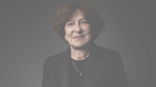Un portrait rapproche d'une femme souriante aux cheveux bruns courts, vetue d'un costume sombre et d'un collier delicat, sur un fond sombre.