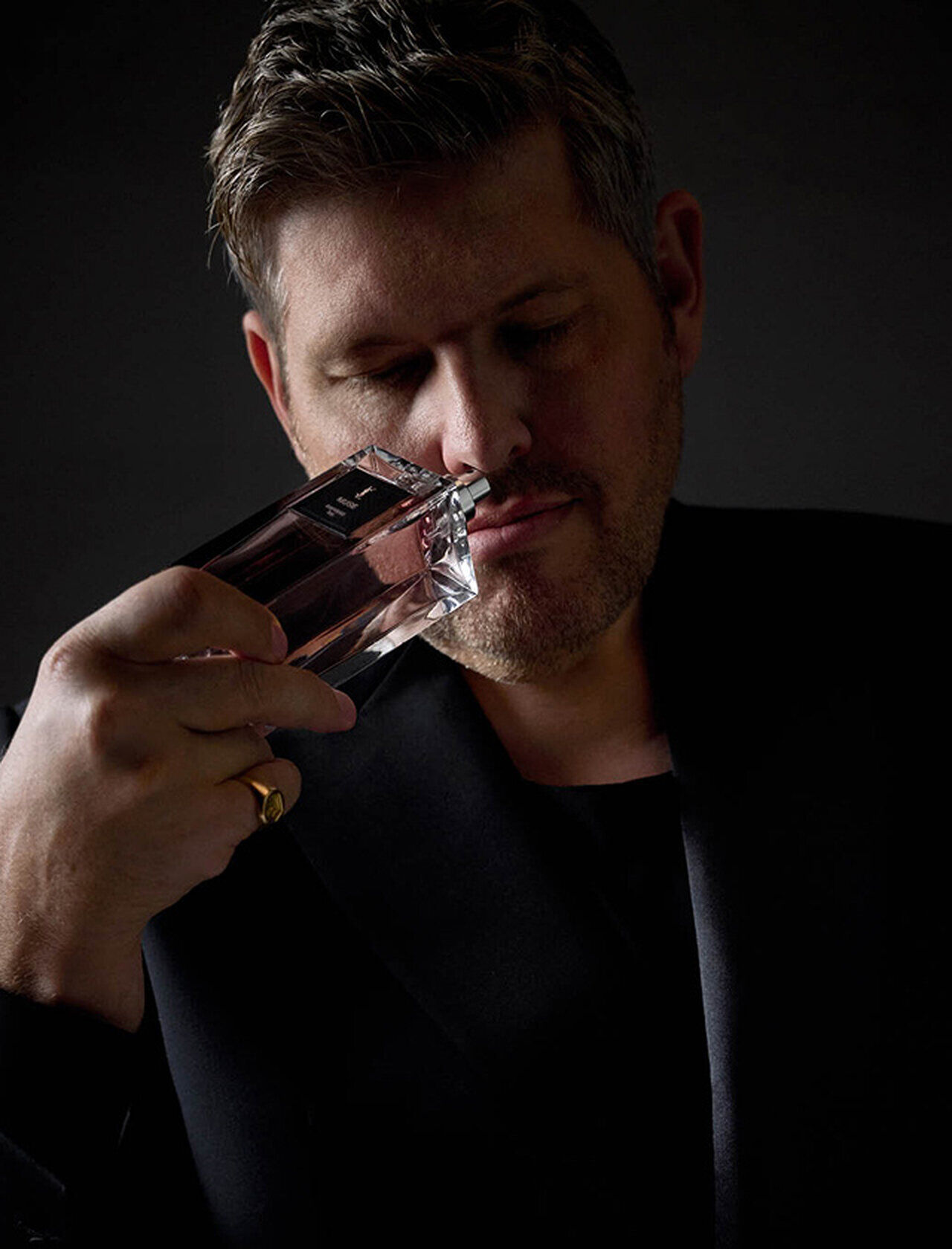 Un homme en haut sombre tient une bouteille de parfum a son nez, inhalant profondement son parfum.