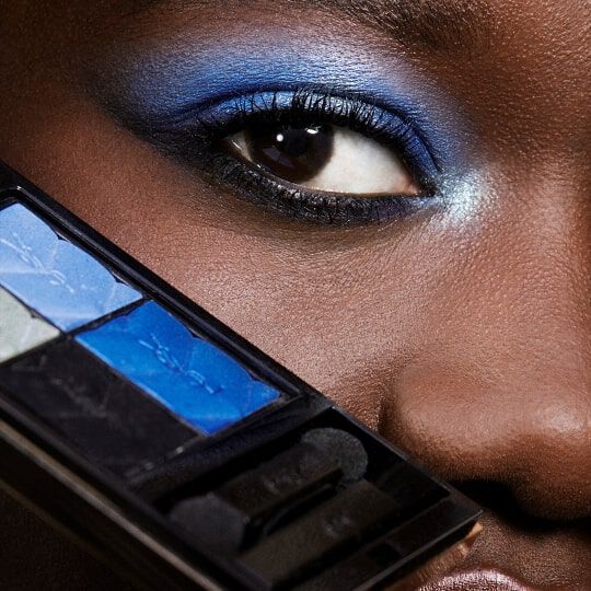 Close-up of an eye featuring bold blue eyeshadow, with a makeup palette displaying various shades of blue held in the foreground.