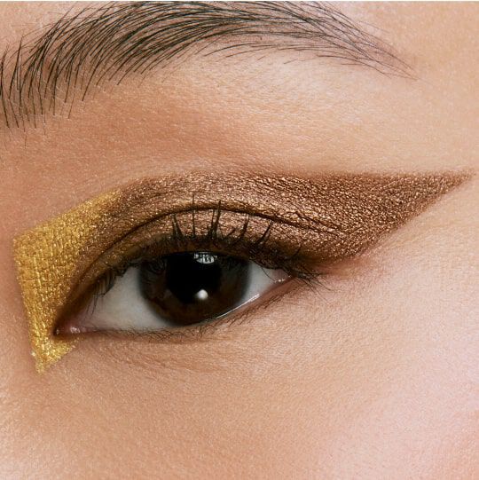 Close-up of an eye with a dramatic makeup look featuring a gold and bronze eyeshadow. The eyeshadow is shaped into a sharp, geometric design emphasizing the eyelid.