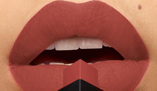 Close-up of a pair of lips wearing a matte lipstick shade, with a lipstick bullet positioned in front, featuring a bold, geometric design.