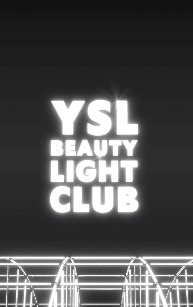 Een brede, donkere opname onthult het "YSL BEAUTY LIGHT CLUB"-logo in helderwitte letters, zwevend in het midden van een zwarte ruimte. Onder het logo creeert een complexe structuur van metalen staven of gloeiende witte neonlichten een raster- of podiumeffect, wat een lichtinstallatie of een futuristische clubomgeving suggereert. Het geheel straalt een moderne en chique sfeer uit.
