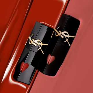 A close-up of two luxurious lipstick tubes in black packaging with gold accents, placed on a vibrant red surface. One tube has a heart-shaped cutout at the base.
