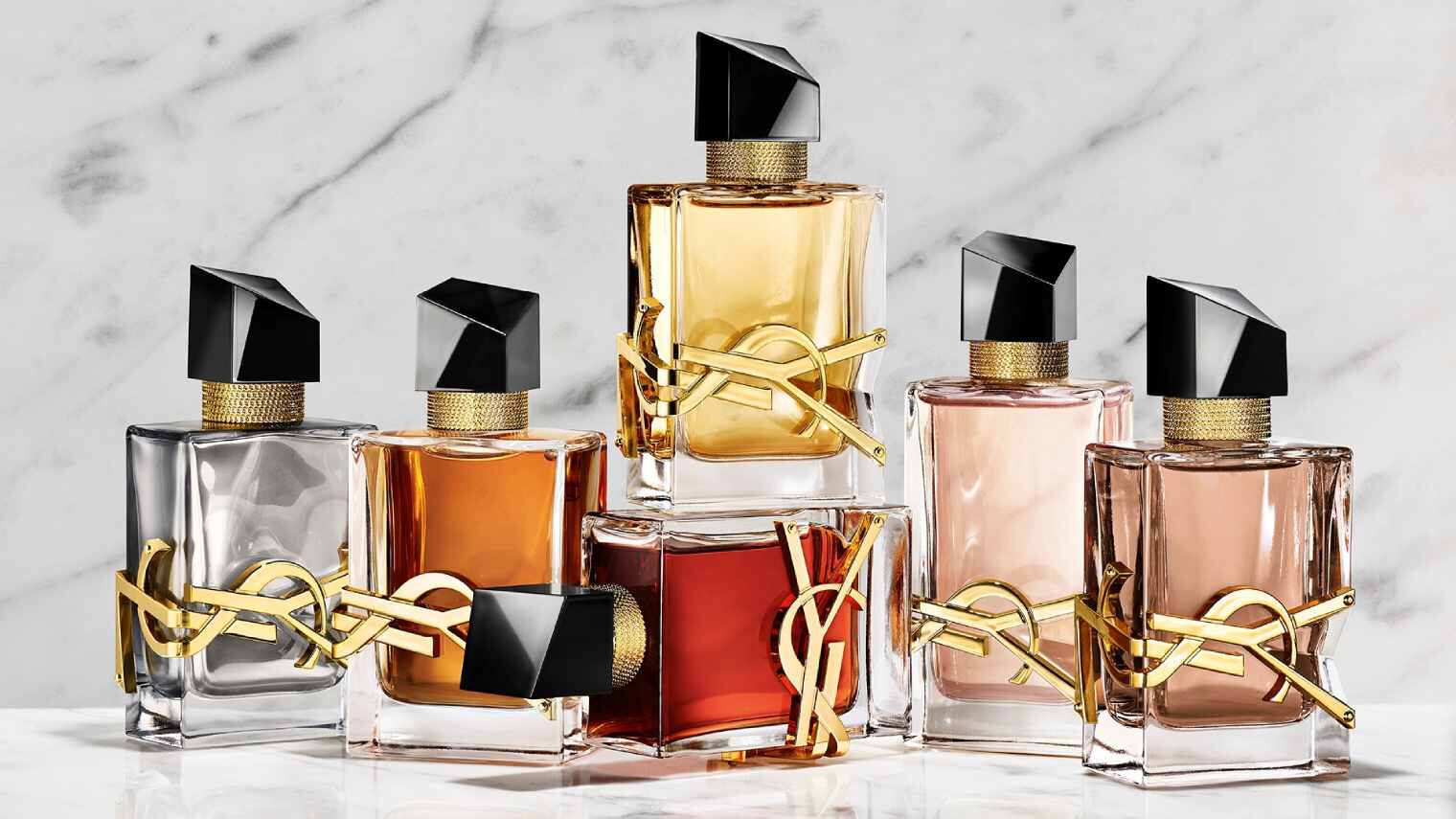 Une main tient un flacon de parfum YSL rouge a cote de deux autres fragrances sur un socle en pierre.