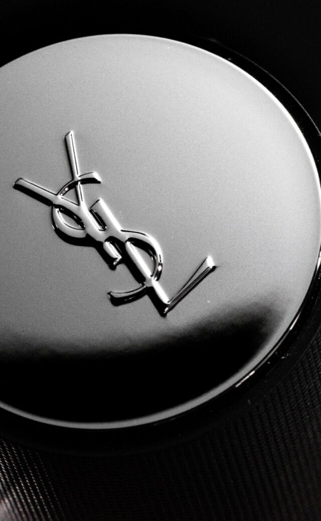 A close-up of a YSL perfume bottle cap, featuring the iconic YSL logo on a shiny, metallic surface. The background is dark, emphasizing the sleek design of the cap.