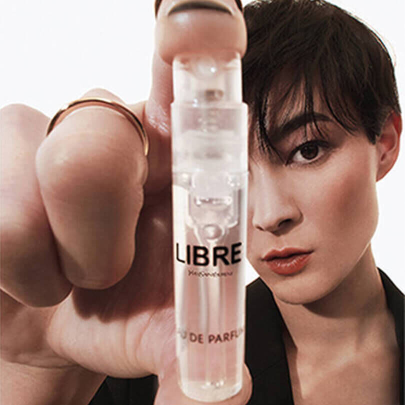 A person with short dark hair holds up a small, clear perfume sample vial of "LIBRE Yves Saint Laurent Eau de Parfum." Their face, with a direct gaze, is partially visible behind their hand and the vial.