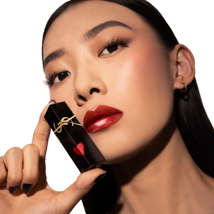 A model with long black hair holds a lipstick tube close to her face, showcasing a bold red lip color with a heart-shaped design on the lipstick packaging. The background is white, emphasizing her makeup look.