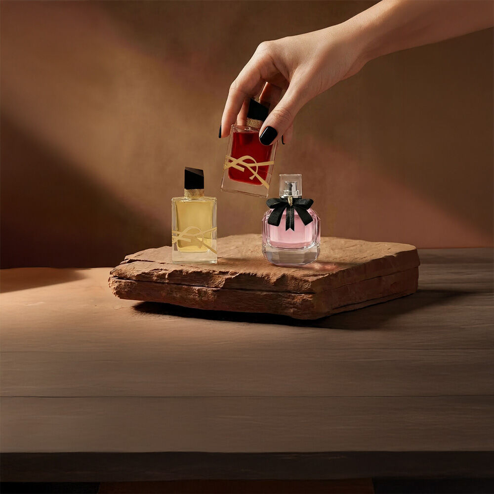 Une main tient un flacon de parfum YSL rouge a cote de deux autres fragrances sur un socle en pierre.