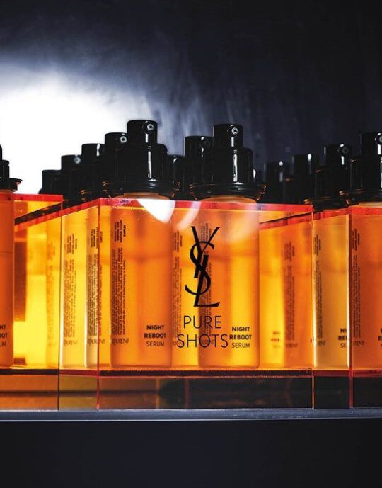 Rij van amberkleurige Yves Saint Laurent Pure Shots Night Reboot Serumflessen met zwarte pompdopjes. Het YSL-logo is prominent aanwezig op elke fles, die op een reflecterend oppervlak staat tegen een donkere, rokerige achtergrond, wat de productlijn benadrukt.