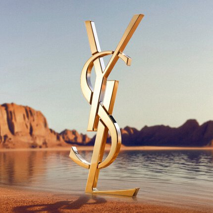 Un grand logo YSL (Yves Saint Laurent) dore en 3D pose sur un rivage desertique. Le monogramme dore poli reflete la lumiere du soleil et se trouve devant une etendue d'eau calme, avec des formations rocheuses rouges spectaculaires et des montagnes desertiques a l'arriere-plan sous un ciel degage.
