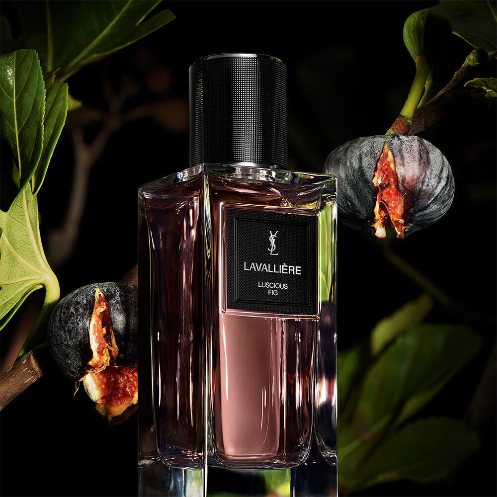 Une bouteille de parfum teintee de violet etiquetee "LAVALLIERE LUSCIOUS FIG" de YSL, avec des branches de figuier et des figues mures et ouvertes.