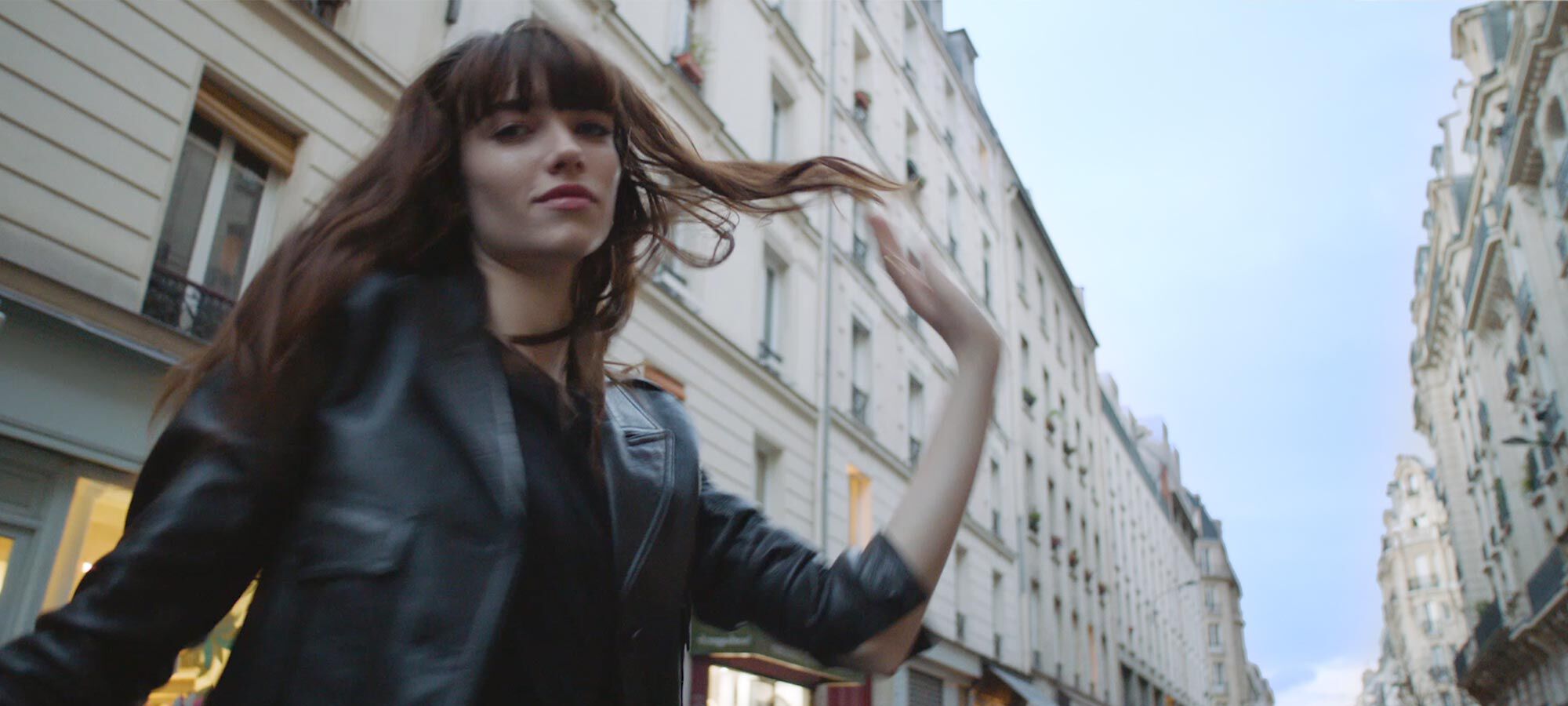 A young woman in a black leather jacket twirls in a Parisian street, with classic white buildings in the background.