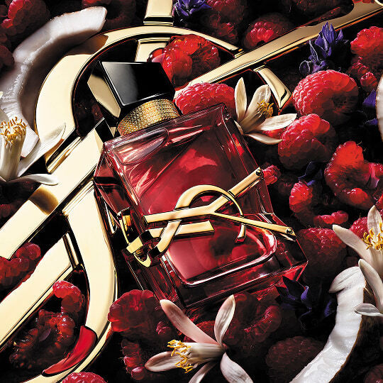 Le flacon rose de Libre Berry Crush d'Yves Saint Laurent entoure de framboises, de fleurs d'oranger blanches et de tranches de noix de coco sur un fond de logo dore.