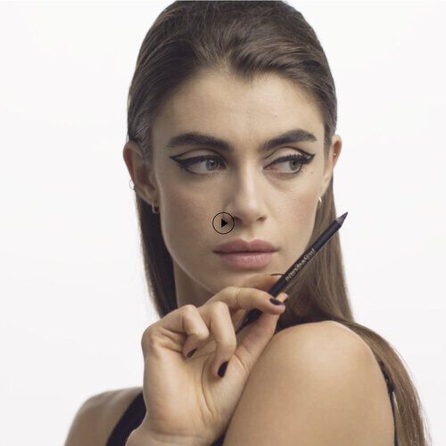 A close-up of a model with dramatic winged eyeliner, holding an eyeliner pencil, and looking thoughtfully to the side. The background is plain white.