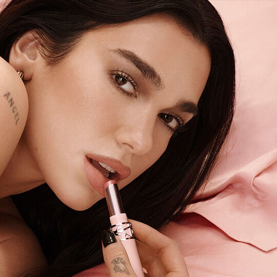 Dua Lipa appliquant un rouge a levres YSL sur fond rose poudre.​