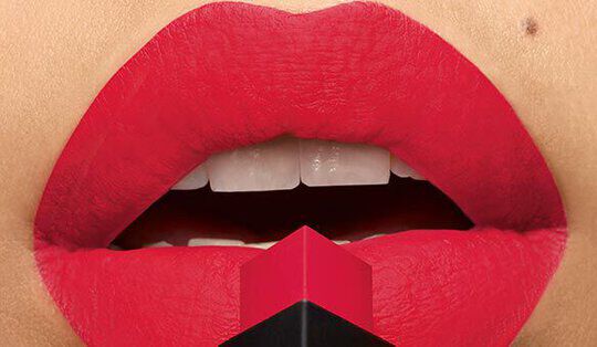 Close-up of a pair of bold red lips with a matte finish, partially holding a red and black lipstick tube near the center.