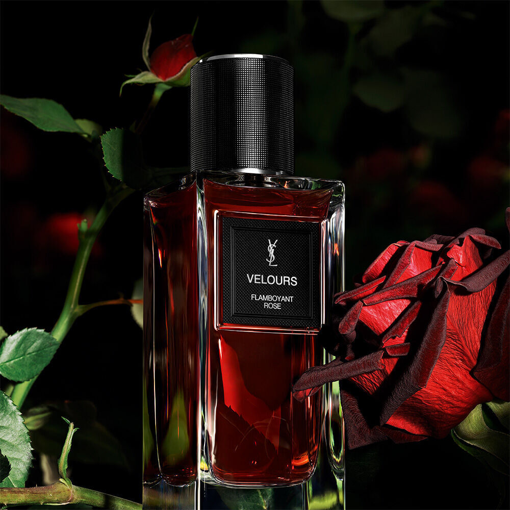 Une bouteille de parfum rouge fonce etiquetee "VELOURS FLAMBOYANT ROSE" de YSL, avec une rose rouge eclatante en arriere-plan flou.