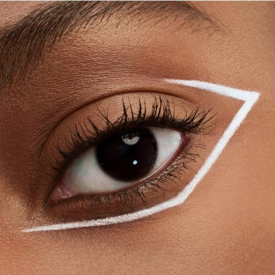 Close-up of an eye with dark lashes and a bold white geometric eyeliner design around it. The skin tone is warm and the background is softly blurred.