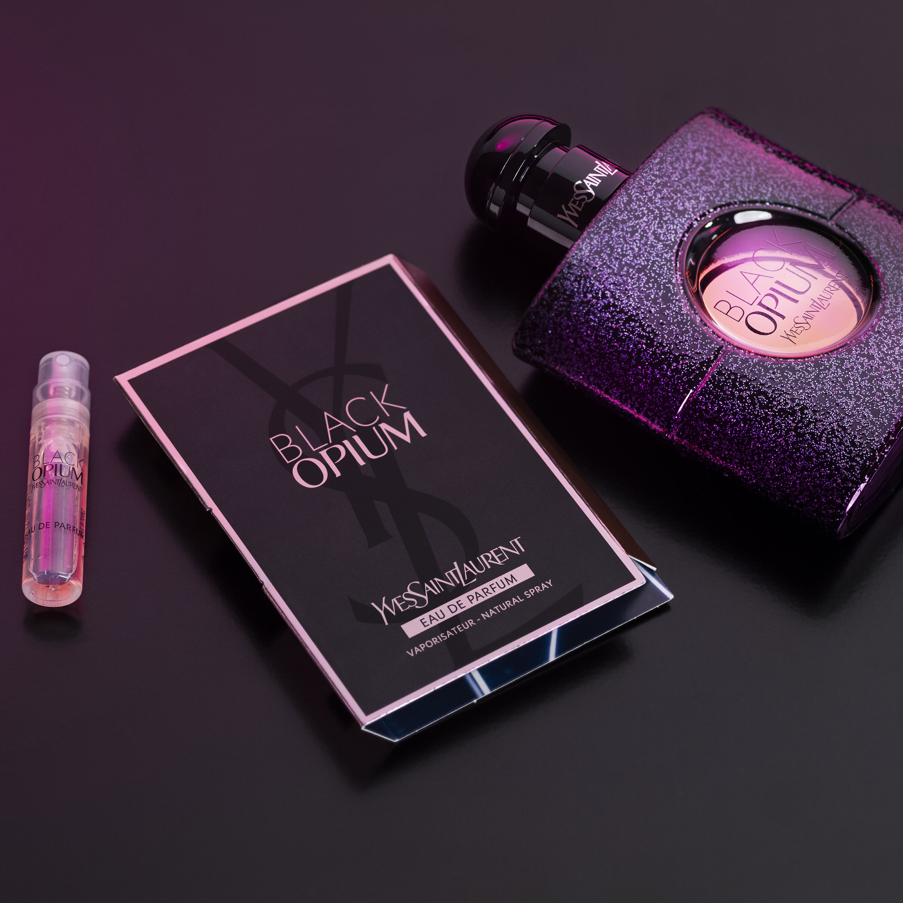 A sparkling bottle of Yves Saint Laurent's Black Opium perfume alongside a sample vial and a promotional card, all set against a dark background.