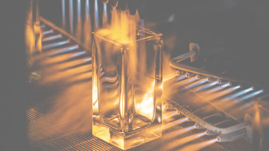 Une bouteille de parfum rectangulaire transparente est montree en train d'etre chauffee par des flammes, probablement dans le cadre d'un processus de fabrication ou de traitement du verre.