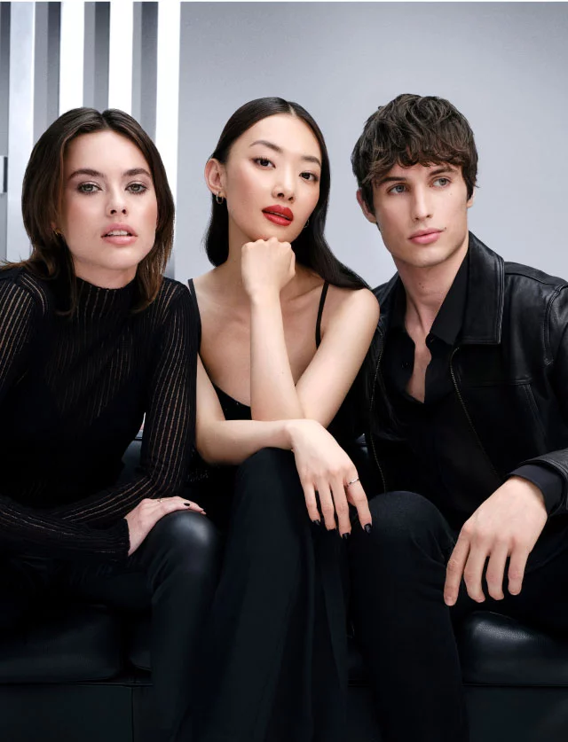Trois jeunes modeles elegants vetus de noir assis sur un canape en cuir noir sur un fond de studio gris.