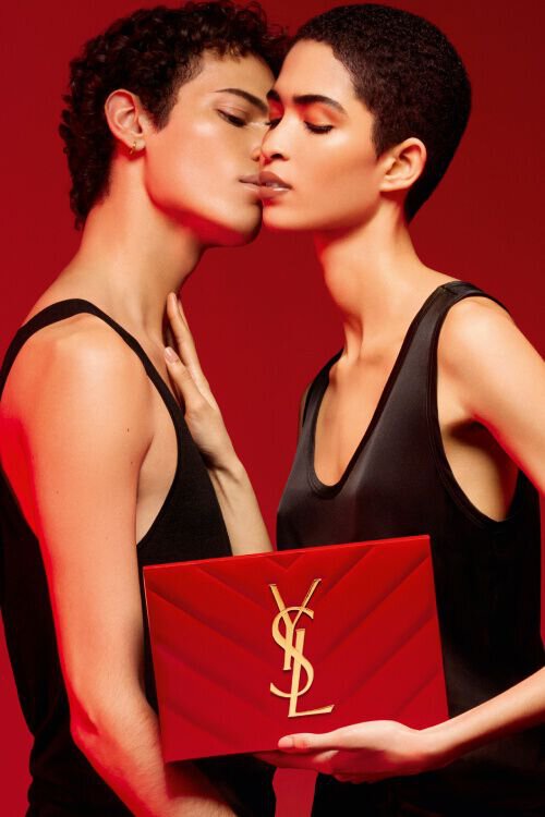 Twee modellen staan dicht bij elkaar met gesloten ogen, een houdt een rode YSL-verpakking vast, tegen een dieprode achtergrond.