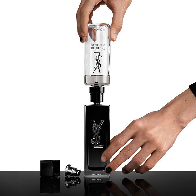 Des mains rechargent un flacon de parfum noir Yves Saint Laurent (YSL) avec une cartouche de recharge transparente indiquant "THE REFILL LA RECHARGE."