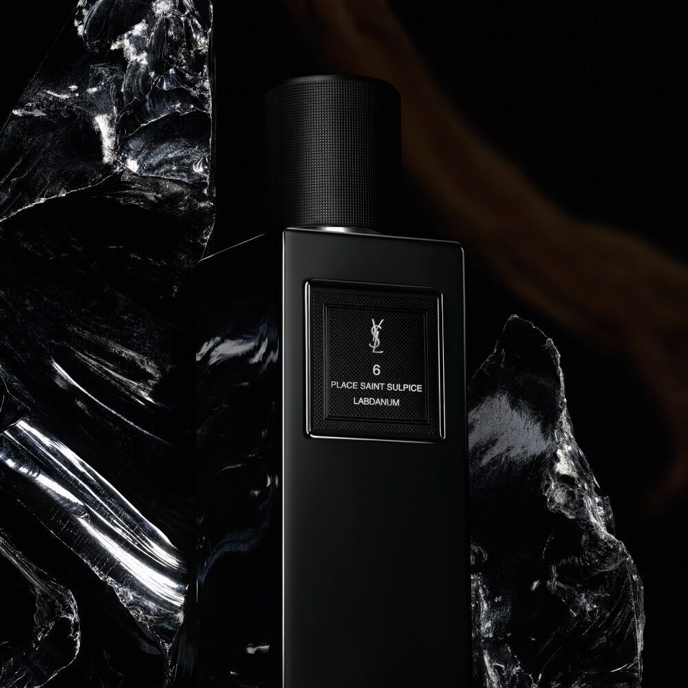 Une elegante bouteille de parfum noire et rectangulaire etiquetee "6 PLACE SAINT SULPICE LABDANUM" de YSL, placee au milieu de superbes pieces de pierre marbree noire brillante.