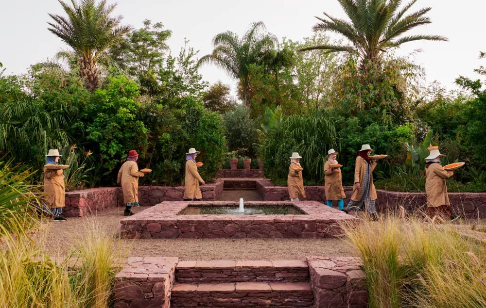A group of people in matching outfits and hats walking in a garden, carrying circular trays. A fountain is in the center, surrounded by lush greenery. The "Ourika Community Gardens" logo is prominently displayed in the foreground.