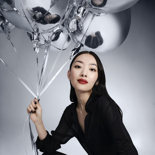 Een vrouw met lang donker haar, gekleed in een zwart overhemd, houdt zilveren ballonnen vast en kijkt recht in de camera. Ze draagt rode lippenstift.