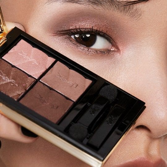 A close-up of a model's eye and hand holding a compact eyeshadow palette with shades of brown and pink.