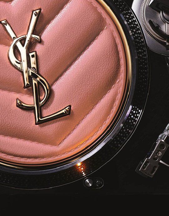 Close-up van een roze, doorgestikt Yves Saint Laurent compact of accessoire met een gouden YSL-logo in het midden. Het item ligt op de metalen, getextureerde rand van een platenspeler, waardoor luxueuze schoonheid wordt gecombineerd met een op muziek geinspireerde esthetiek.