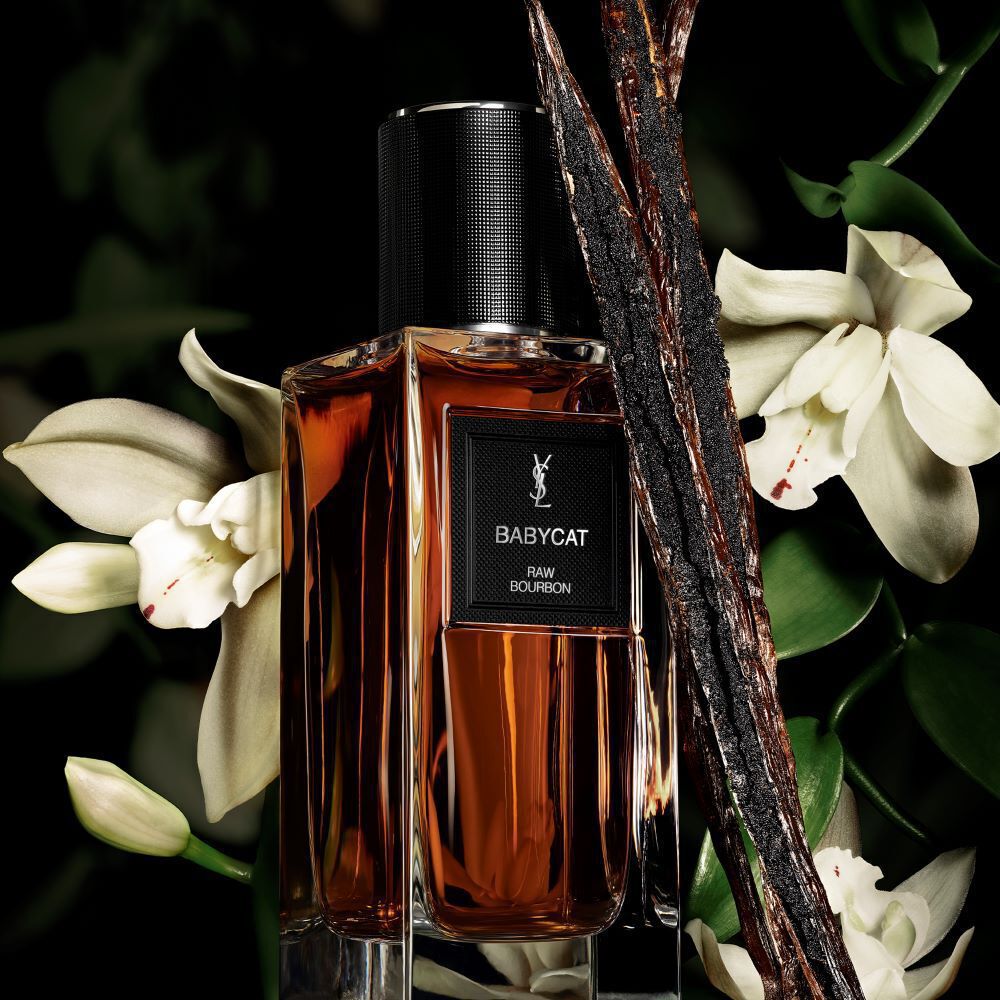 Une bouteille de parfum couleur ambre etiquetee "BABYCAT RAW BOURBON" de YSL, ornee de gousses de vanille et de fleurs d'orchidees blanches.