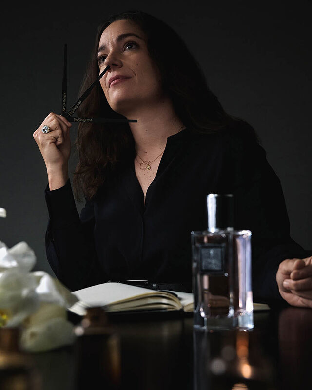 Une femme en chemise noire tient une touche a parfum, levant les yeux pensivement, avec un carnet, une bouteille de parfum et des fleurs sur la table.