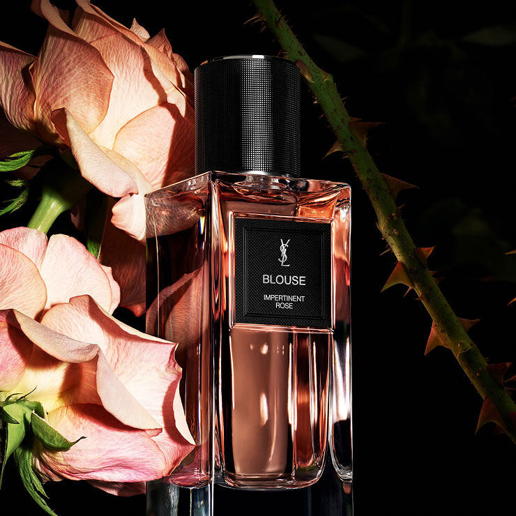 Une bouteille de parfum rectangulaire etiquetee "BLOUSE IMPERTINENT ROSE" de YSL, entouree de roses roses pales et de tiges de roses epineuses.