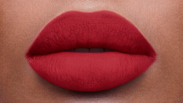 Close-up of a pair of full lips coated in a matte red lipstick against a smooth, beige background.