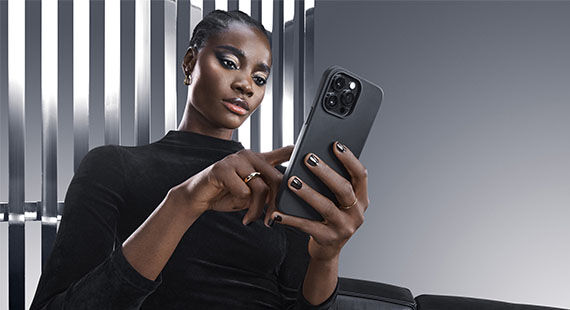 A model with braided hair is sitting on a sleek couch, focused on using a smartphone, while dressed in a black top against a modern, striking background.