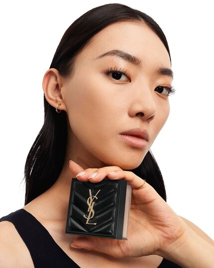 Une femme tient un poudrier carre noir Yves Saint Laurent pres de son visage.