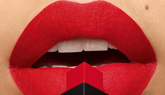 Close-up of bold red lips with a matte finish, holding a red and black lipstick bullet in front of them.