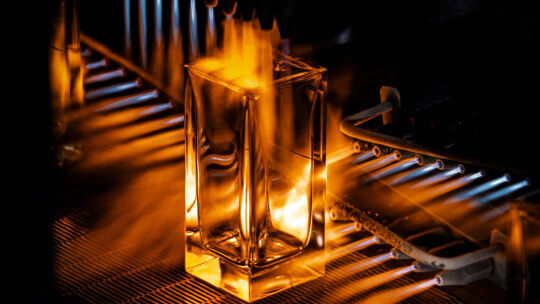 Une bouteille de parfum rectangulaire transparente est montree en train d'etre chauffee par des flammes, probablement dans le cadre d'un processus de fabrication ou de traitement du verre.