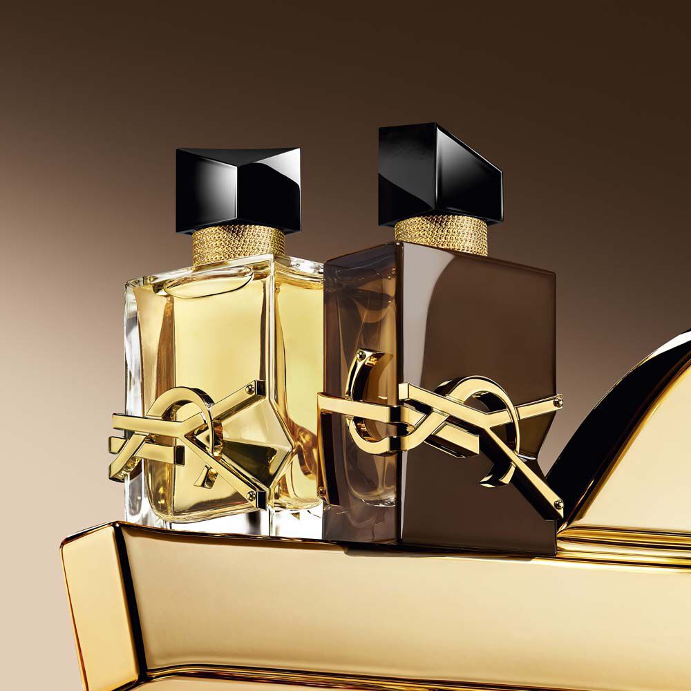 Glazed brown Libre Santal Couture bottle and Libre Eau de Parfum bottle reflecting light, placed on a large golden YSL Cassandre.