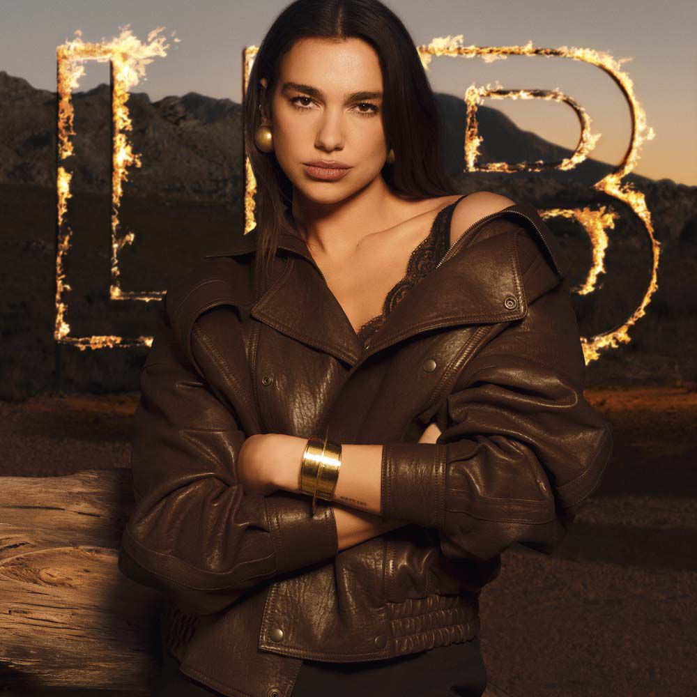 Dua Lipa in a brown leather jacket with large flaming LIBRE letters in a raw desert landscape.